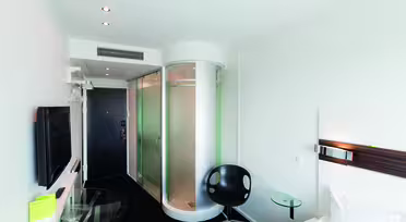 Modernt hotellrum med elegant design, med en säng, en platt-TV på väggen, en rund stol, ett sidobord med glasskiva och en distinkt cylindrisk duschvägg i glas nära entrédörren.
