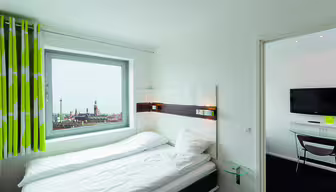 Minimalistiskt hotellrum med en snyggt bäddad säng, vita sängkläder och grönrandiga gardiner. Ett fönster visar en stadsbild. En monterad TV finns i det intilliggande rummet, synlig genom en öppen dörr. Väggar och möbler är övervägande vita.