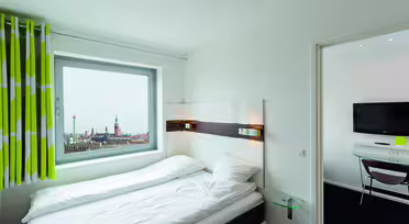 Minimalistiskt hotellrum med en snyggt bäddad säng, vita sängkläder och grönrandiga gardiner. Ett fönster visar en stadsbild. En monterad TV finns i det intilliggande rummet, synlig genom en öppen dörr. Väggar och möbler är övervägande vita.