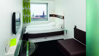 Modernt hotellrum med dubbelsäng, väggmonterad TV och skrivbord i glas. Ett stort fönster erbjuder utsikt över stadsbilden med två framträdande torn. Brun soffa och grönmönstrade gardiner kompletterar den minimalistiska inredningen.