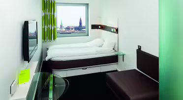 Modernt hotellrum med dubbelsäng, väggmonterad TV och skrivbord i glas. Ett stort fönster erbjuder utsikt över stadsbilden med två framträdande torn. Brun soffa och grönmönstrade gardiner kompletterar den minimalistiska inredningen.