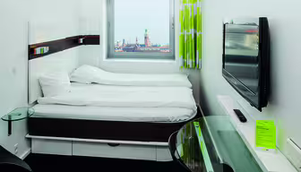 Modernt hotellrum med en dubbelsäng under ett fönster som visar en stadsbild. En platt-TV är monterad på väggen och det finns ett skrivbord i glas med en telefon. Rummet har gröna och vita gardiner.