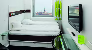 Modernt hotellrum med en dubbelsäng under ett fönster som visar en stadsbild. En platt-TV är monterad på väggen och det finns ett skrivbord i glas med en telefon. Rummet har gröna och vita gardiner.