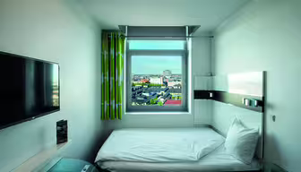 Kompakt hotellrum med en enkelsäng. Rummet har en väggmonterad TV, ett fönster med stadsutsikt, grönmönstrade gardiner, ett litet bord och minimalistisk modern inredning. Sängen är snyggt gjord med vita sängkläder.