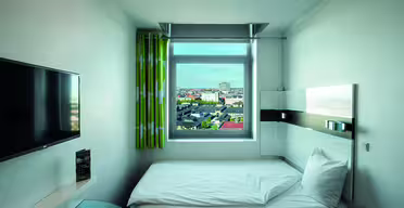 Kompakt hotellrum med en enkelsäng. Rummet har en väggmonterad TV, ett fönster med stadsutsikt, grönmönstrade gardiner, ett litet bord och minimalistisk modern inredning. Sängen är snyggt gjord med vita sängkläder.