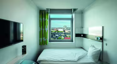 Kompakt hotellrum med en enkelsäng. Rummet har en väggmonterad TV, ett fönster med stadsutsikt, grönmönstrade gardiner, ett litet bord och minimalistisk modern inredning. Sängen är snyggt gjord med vita sängkläder.