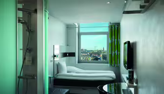 Kompakt modernt hotellrum med en vit säng bredvid ett stort fönster, med utsikt över stadsbilden. Rummet har en väggmonterad TV, ett litet runt bord med stol och en inglasad dusch. Grönrandiga gardiner sätter färg på den snygga vita inredningen.