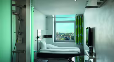 Kompakt modernt hotellrum med en vit säng bredvid ett stort fönster, med utsikt över stadsbilden. Rummet har en väggmonterad TV, ett litet runt bord med stol och en inglasad dusch. Grönrandiga gardiner sätter färg på den snygga vita inredningen.
