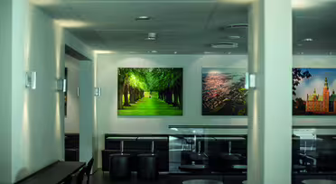 En modern kaféinteriör med minimalistisk design, med svarta bord och pallar. På väggen finns stora inramade foton av en trädkantad stig och en stadsvy från luften, vilket ger utrymmet en levande och naturskön touch.