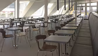 Modern cafeteria med långa sittplatser längs en vägg av fönster. Rummet har rader av bord med metall- och trästolar, och parallellt med borden löper en skinnbänk. Naturligt ljus strömmar genom stora, vinklade fönster.