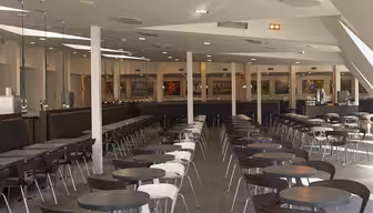 Bild av en tom cafeteria med rader av bord och stolar. Rummet är rymligt, med högt i tak och modern belysning. Vita och mörkbruna stolar paras ihop med matchande runda bord på ett grått klinkergolv. Stora fönster släpper in naturligt ljus.