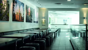 En modern, tom cafeteria med en rad fyrkantiga bord och runda pallar till vänster. Väggarna pryds av inramade landskapsfoton. Solljus strömmar in genom stora fönster och skapar en ljus och luftig atmosfär.