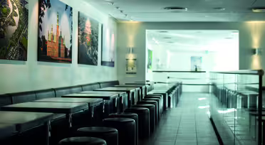 En modern, tom cafeteria med en rad fyrkantiga bord och runda pallar till vänster. Väggarna pryds av inramade landskapsfoton. Solljus strömmar in genom stora fönster och skapar en ljus och luftig atmosfär.