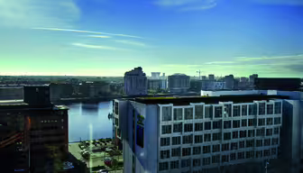 Stadsbild av ett hamnområde med moderna byggnader under en klarblå himmel. En vattenmassa är i centrum, omgiven av höghus och några parkerade bilar synliga längst ner.