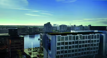 Stadsbild av ett hamnområde med moderna byggnader under en klarblå himmel. En vattenmassa är i centrum, omgiven av höghus och några parkerade bilar synliga längst ner.