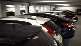 Rader av parkerade bilar i ett svagt upplyst inomhusparkeringsgarage, med fordon i olika riktningar. Scenen visar garagets betongtak och väggar, vilket skapar en typisk urban parkeringsmiljö.