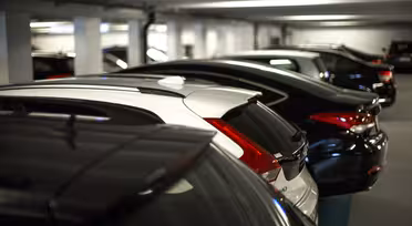 Rader av parkerade bilar i ett svagt upplyst inomhusparkeringsgarage, med fordon i olika riktningar. Scenen visar garagets betongtak och väggar, vilket skapar en typisk urban parkeringsmiljö.