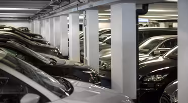 Ett svagt upplyst underjordiskt parkeringsgarage fyllt med parkerade bilar. Fordonen är placerade tätt intill varandra, med synliga pelare som stödjer taket. Scenen verkar organiserad och lugn.