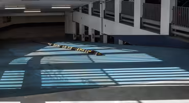 Ljus och skuggor skapar randiga mönster på golvet i ett tomt parkeringsgarage. Ett långt farthinder går över betongytan och utrymmet är upplyst av lysrör. Den övergripande tonen är cool och industriell.