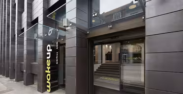 Modern hotellentré med mörk tegelfasad och stor glastak. Gul "wakeup"-logotyp nära de öppna skjutdörrarna i glas. Steg inuti leder till receptionen. Urban miljö synlig genom glasreflektion.