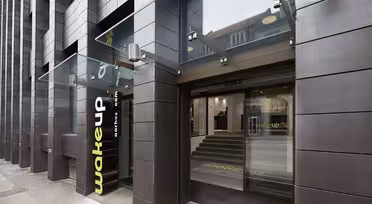Modern hotellentré med mörk tegelfasad och stor glastak. Gul "wakeup"-logotyp nära de öppna skjutdörrarna i glas. Steg inuti leder till receptionen. Urban miljö synlig genom glasreflektion.