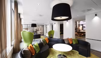 Modern loungedel med en rund svart soffa med färgglada kuddar runt ett vitt bord på en grön matta. Gröna wingback-stolar och stora svarta taklampor bidrar till den moderna designen. Bruna gardiner ramar in stora fönster och lyser upp utrymmet.