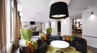 Modern loungedel med en rund svart soffa med färgglada kuddar runt ett vitt bord på en grön matta. Gröna wingback-stolar och stora svarta taklampor bidrar till den moderna designen. Bruna gardiner ramar in stora fönster och lyser upp utrymmet.