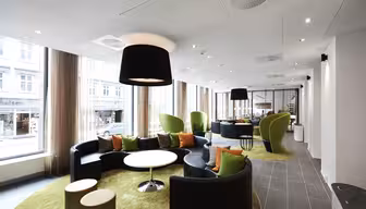 Modern lounge med runda svarta soffor, gröna kuddar och vita bord på gröna mattor. Stora fönster ger naturligt ljus. Snygga svartvita belysningsarmaturer hänger i taket och utrymmet har utsikt över en stadsgata.