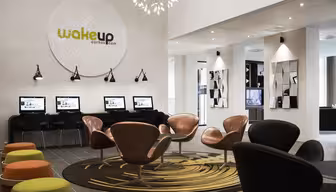 Modern hotellobby med en rad datorer mot väggen under en stor cirkulär skylt där det står "wakeup aarhus.com." Snygga accenter inkluderar bruna och svarta stolar, färgglada pallar, en mönstrad matta och väggspeglar. Ljus belysning och modern design.
