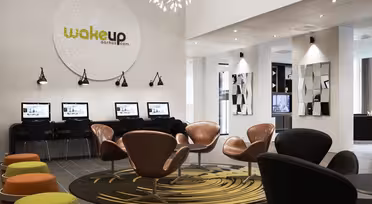 Modern hotellobby med en rad datorer mot väggen under en stor cirkulär skylt där det står "wakeup aarhus.com." Snygga accenter inkluderar bruna och svarta stolar, färgglada pallar, en mönstrad matta och väggspeglar. Ljus belysning och modern design.