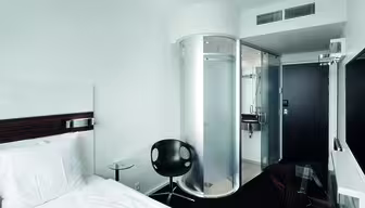 Modernt hotellrum med en enkelsäng, vita sängkläder och ett litet runt glasbord med en svart stol. En cylindrisk glasdusch och ett kompakt badrum finns bredvid en svart dörr. En stor platt-TV är monterad på väggen mittemot sängen.