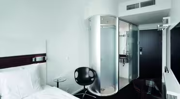 Modernt hotellrum med en enkelsäng, vita sängkläder och ett litet runt glasbord med en svart stol. En cylindrisk glasdusch och ett kompakt badrum finns bredvid en svart dörr. En stor platt-TV är monterad på väggen mittemot sängen.