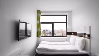Ett litet, modernt hotellrum med en snyggt bäddad säng med vita sängkläder, en väggmonterad TV och ett fönster med utsikt över stadens byggnader. Väggarna är vita och det finns en ljusgrön gardin på fönstret.
