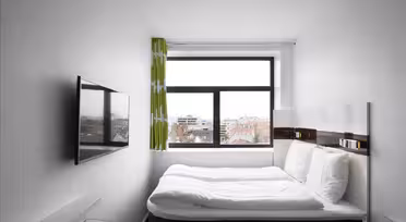 Ett litet, modernt hotellrum med en snyggt bäddad säng med vita sängkläder, en väggmonterad TV och ett fönster med utsikt över stadens byggnader. Väggarna är vita och det finns en ljusgrön gardin på fönstret.