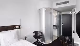 Modernt hotellrum med vit säng, svart stol och skrivbord i glas. En unik cylindrisk dusch med frostat glas står nära entrén. Rummet har minimalistisk inredning och vita väggar, vilket skapar en ren och modern atmosfär.