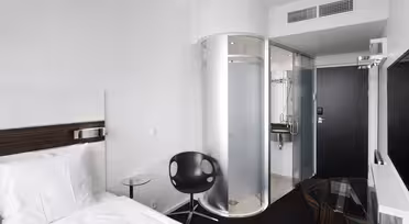 Modernt hotellrum med vit säng, svart stol och skrivbord i glas. En unik cylindrisk dusch med frostat glas står nära entrén. Rummet har minimalistisk inredning och vita väggar, vilket skapar en ren och modern atmosfär.