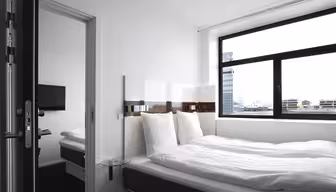 Modernt hotellrum med stor säng, vita sängkläder och mörk sängram. Ett litet sidobord i glas står bredvid sängen. Ett brett fönster erbjuder utsikt över stadens byggnader. En platt-TV är synlig i ett angränsande rum genom en öppen dörr.
