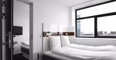 Modernt hotellrum med stor säng, vita sängkläder och mörk sängram. Ett litet sidobord i glas står bredvid sängen. Ett brett fönster erbjuder utsikt över stadens byggnader. En platt-TV är synlig i ett angränsande rum genom en öppen dörr.