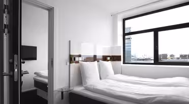Modernt hotellrum med stor säng, vita sängkläder och mörk sängram. Ett litet sidobord i glas står bredvid sängen. Ett brett fönster erbjuder utsikt över stadens byggnader. En platt-TV är synlig i ett angränsande rum genom en öppen dörr.