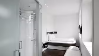 Minimalistiskt hotellrum med en inglasad dusch till vänster och en säng med vita sängkläder mot väggen till höger. Ett litet skrivbord med stol och en väggmonterad TV syns också. Rummet har elegant, modern inredning.