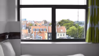 Utsikt från ett fönster som visar en stadsbild med byggnader med röda tak och frodiga träd. I förgrunden finns en del av en säng med vita sängkläder, och grönmönstrade gardiner ramar delvis in fönstret.