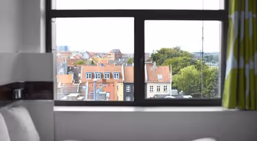 Utsikt från ett fönster som visar en stadsbild med byggnader med röda tak och frodiga träd. I förgrunden finns en del av en säng med vita sängkläder, och grönmönstrade gardiner ramar delvis in fönstret.