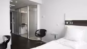Minimalistiskt hotellrum med en snyggt gjord vit säng, mörkt trägolv och en modern stol bredvid ett litet glasbord. Badrummet är omslutet av frostade glasdörrar, med snygg inredning. Ljus belysning och rena linjer förstärker den moderna känslan.