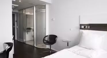 Minimalistiskt hotellrum med en snyggt gjord vit säng, mörkt trägolv och en modern stol bredvid ett litet glasbord. Badrummet är omslutet av frostade glasdörrar, med snygg inredning. Ljus belysning och rena linjer förstärker den moderna känslan.