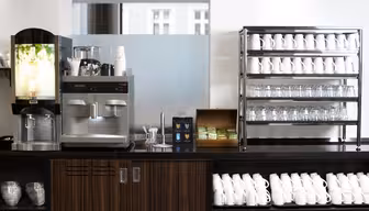 En kaffestation med en automat för varma drycker, en espressomaskin och hyllor fyllda med vita muggar och glas. En trälåda med tepåsar och koppar placeras mellan maskinerna.