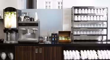 En kaffestation med en automat för varma drycker, en espressomaskin och hyllor fyllda med vita muggar och glas. En trälåda med tepåsar och koppar placeras mellan maskinerna.