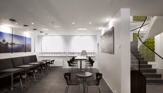 Modern kontorsyta med eleganta svarta sittplatser, runda bord med träytor och minimalistisk design. En spiraltrappa är till höger, och de vita väggarna har konstverk med nautiskt tema med svaga takspotlights för belysning.