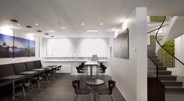 Modern kontorsyta med eleganta svarta sittplatser, runda bord med träytor och minimalistisk design. En spiraltrappa är till höger, och de vita väggarna har konstverk med nautiskt tema med svaga takspotlights för belysning.