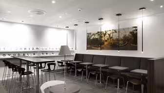 Modern caféinteriör med elegant design. Har höga bord med pallar och en lång monter med små bord. Ett stort foto av en solbelyst kupolträdgård hänger på väggen. Taklampor och en dekorativ bokstavspanel bidrar till den minimalistiska atmosfären.