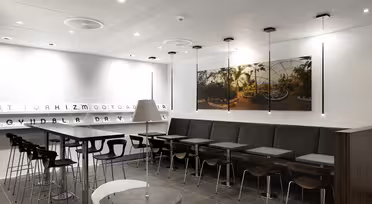Modern caféinteriör med elegant design. Har höga bord med pallar och en lång monter med små bord. Ett stort foto av en solbelyst kupolträdgård hänger på väggen. Taklampor och en dekorativ bokstavspanel bidrar till den minimalistiska atmosfären.